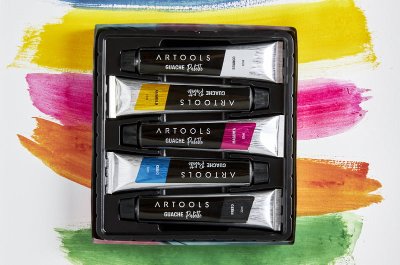 Caixa com cinco bisnagas de tinta guache da Artools em cima de um papel com pinceladas grossas de tintas coloridas 