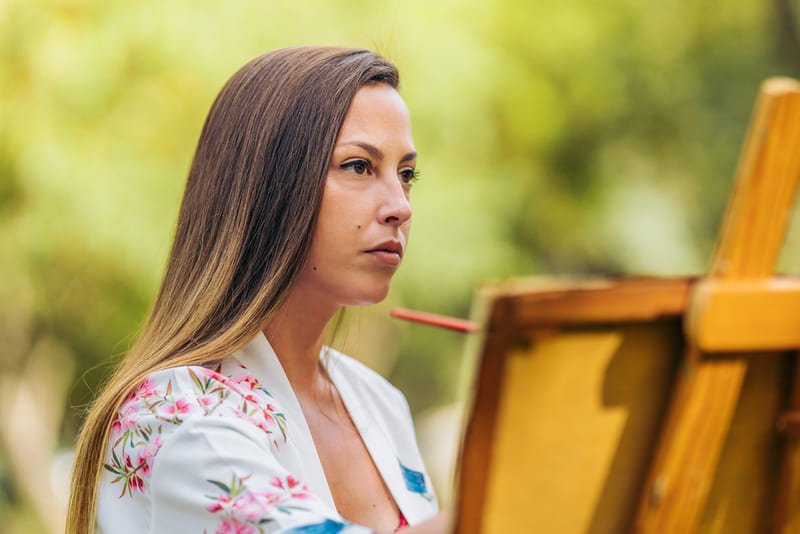 Jovem mulher observando algo e desenhando em tela que está apoiada em um cavalete